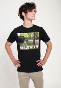 Sort bomulds T-shirt med et stort grafisk print af en gade-scene fra "Halloween" med grønne træer og titlen i gul tekst.