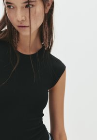 Crna, uska majica s kratkim rukavima i okruglim izrezom, napravljena od rastezljivog materijala. Model nosi perle naušnice, a raspuštena kosa okviruje lice.
