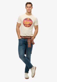 Camiseta de hombre de color crema con estampado gráfico retro, jeans azules y una camisa de cuadros rojos atada a la cintura. Zapatillas blancas con detalles marrones.
