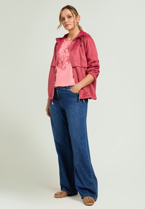 Femme portant une veste à capuche rouge, une chemise fleurie rose, un jean large bleu et des mocassins marron, posant avec une main dans la poche.