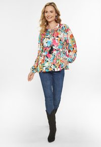 Bunte Bluse mit Blumenmuster, gerafftem Bund, langen Ärmeln und Quasten-Details. Kombiniert mit blauen Jeans und schwarzen Wildleder-Cowboy-Stiefeln.