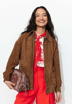 Femme aux longs cheveux foncés souriante, portant une veste en daim marron, un foulard bandana rouge, un t-shirt blanc graphique, un pantalon rouge, tenant un sac marron à motif paisley.