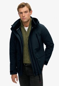 Marineblauwe waterdichte jas met een capuchon, voorzien van voorzakken, verstelbare manchetten en een ritsluiting, gedragen over een olijfgroene kabelgebreide trui.