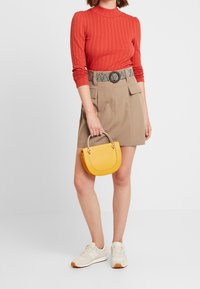Red ribbed long-sleeve top, beige high-waisted shorts with pockets, black and white patterned belt, and a small yellow handbag. White sneakers.