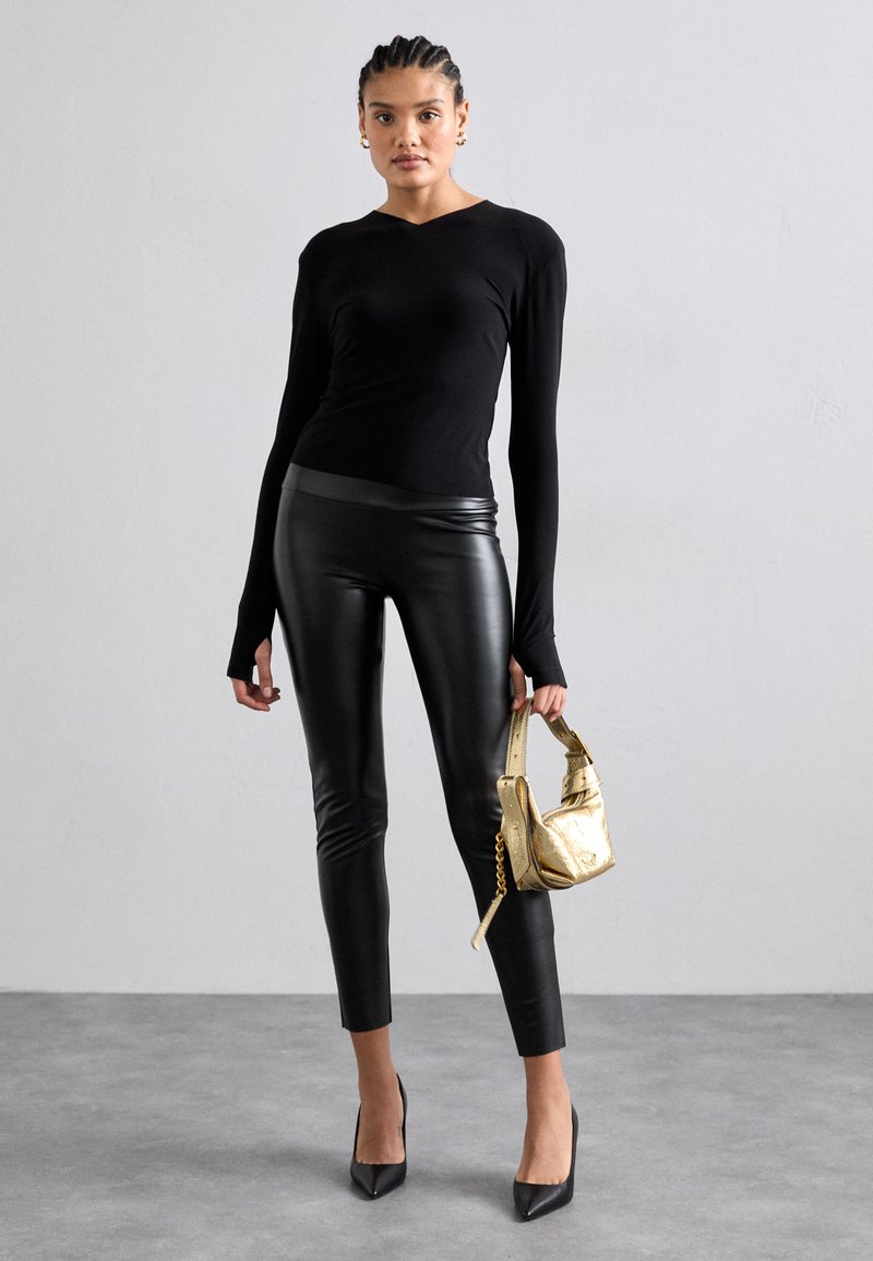 Schwarzes Langarmoberteil, eng anliegende, dehnbare schwarze Kunstlederleggings, spitze schwarze High Heels und eine kleine metallisch goldene Handtasche.