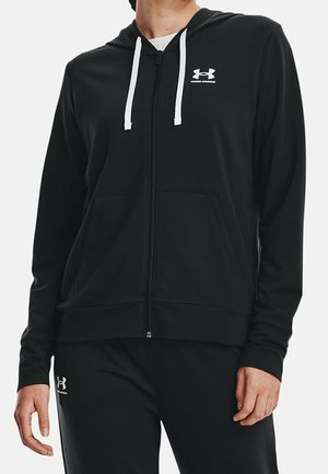 Osoba nosząca czarną bluzę Under Armour na suwak z białymi troczkami oraz dopasowane czarne spodnie z logo na lewej udzie.