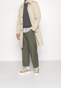 Beige trenchcoat over een donkere trui, gestreept shirt en groene broek. Schoeisel: beige sneakers met witte zolen, gecombineerd met witte sokken.
