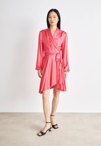 Rosa sateng omslagskjole med lange ermer, en knyttet midje og en asymmetrisk kant. Sammen med svarte sandaler med stropper på en nøytral gulv.