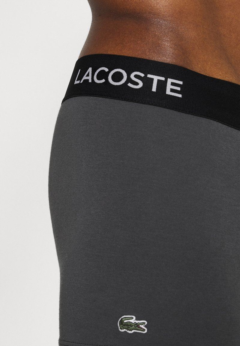 Boxer gris avec une taille noire arborant le texte "LACOSTE" en blanc et un petit logo crocodile vert en bas à gauche.