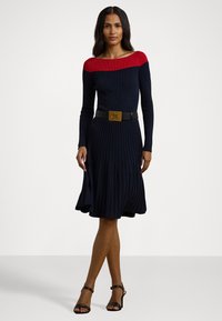 Vestido de malha com riscas em azul-marinho e vermelho, com um corpete ajustado, saia rodada e plissada, e um cinto largo preto com uma fivela dourada em forma de âncora.