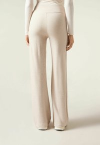 Beige geribde wijde broek met een hoge taille, zachte textuur en geen zichtbare patronen. De broek loopt tot de vloer en is gecombineerd met witte schoenen.