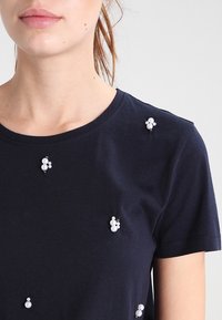 Námořnické bavlněné tričko s dekorativními perlami různých velikostí uspořádanými asymetricky na přední straně. Krátké rukávy, kulatý výstřih.