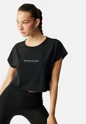 Jonge vrouw met gevlochten haar, zilveren hoepeloorbellen en een zwarte cropped top met de tekst "ONE STEP UP LEVEL" in het wit.