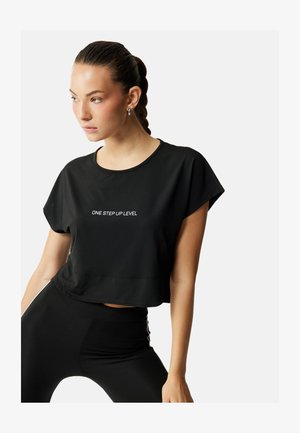 Jonge vrouw met gevlochten haar, zilveren hoepeloorbellen en een zwarte cropped top met de tekst "ONE STEP UP LEVEL" in het wit.