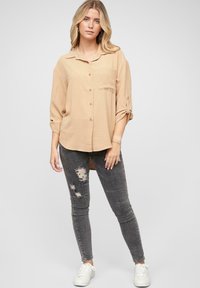 Bluză bej cu nasturi, dotată cu un buzunar, de croială lejeră, având mâneci desfăcute. Purtați-o cu blugi skinny gri, decolorati și rupți, și adidași albi.