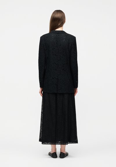 Γυναίκα που στέκεται πλάτη προς τα εμπρός φορώντας ένα μακρύ μαύρο δαντελένιο σακάκι και ταιριαστή φούστα μέχρι τον αστράγαλο με μαύρα επίπεδα παπούτσια.