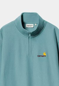 Sudadera azul claro con cuello de media cremallera, que presenta un logo bordado en amarillo y burdeos. Tejido suave y texturizado.