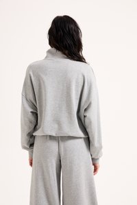 Γκρι φωτεινή μπλούζα με ψηλό γιακά και χαμηλούς ώμους, σε συνδυασμό με παρόμοιες χαλαρές παντελόνες, που αναδεικνύουν μια απαλή, υφασμένη ύλη.