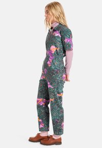 Jumpsuit i mørkegrønn med et livlig blomstermønster i rosa, lilla og oransje. Har glidelås foran og korte ermer.