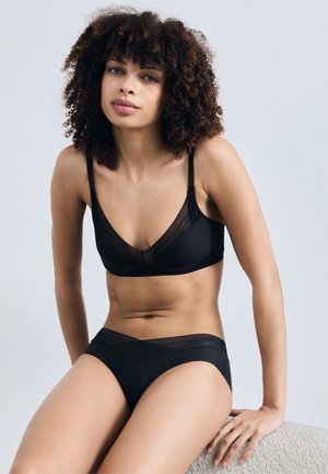 Kobieta o kręconych włosach, ubrana w czarny biustonosz i dopasowaną bieliznę, siedząca i patrząca przed siebie na jednolitym jasnoszarym tle.