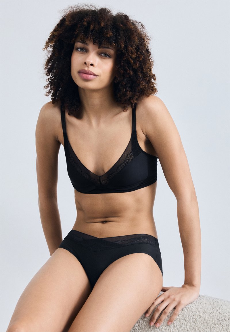 Γυναίκα με σγουρά μαλλιά που φοράει μαύρο σουτιέν και αντίστοιχο εσώρουχο, καθισμένη και κοιτάζει μπροστά με φόντο ένα απλό ανοιχτό γκρι.