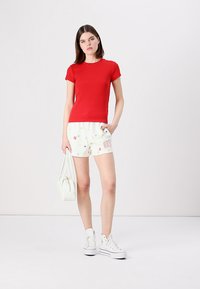 Rød bomulds T-shirt parret med hvide shorts med blomstermønster og "GARD" tekst. Modellen holder en hvid håndtaske og har hvide sneakers på.