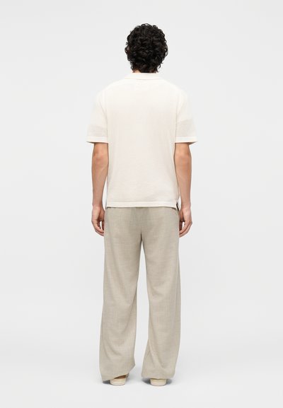 Άνδρας με σγουρά μαλλιά, φορώντας λευκό υφασμάτινο κοντομάνικο πουκάμισο και μπεζ παντελόνια με φαρδιά πόδια, στέκεται με την πλάτη προς τη λευκή φόντο.