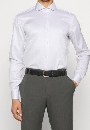 Mężczyzna ubrany w dopasowaną białą koszulę włożoną do szarych spodni, z czarnym skórzanym paskiem, jedna ręka w kieszeni, na jednolitym tle.