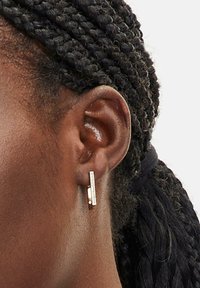 Goldfarbene rechteckige Ohrringe mit einem eleganten, modernen Design. Sie verfügen über eine strukturierte Oberfläche und einen minimalistischen Stil und werden zu einer geflochtenen Frisur getragen.
