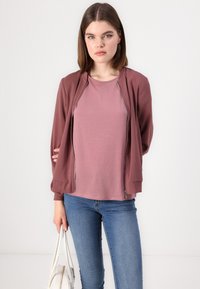 Hindbær zip-up bomberjakke over en lilac T-shirt, kombinert med blå jeans. Modellen holder en hvit håndveske, og står mot en lys bakgrunn.