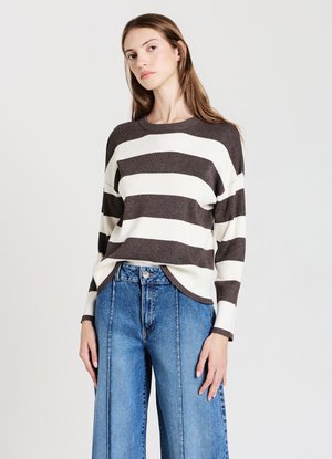 Junge Frau mit langen braunen Haaren, die einen braun-weiß gestreiften Pullover und hoch taillierte blaue Jeans trägt, steht vor einem schlichten Hintergrund.