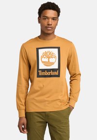 Long-sleeve, mustard yellow shirt made of cotton featuring a black-bordered white logo with a tree and "Timberland" text at the bottom.