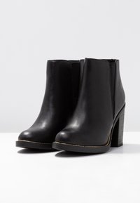 Botas negras hasta el tobillo hechas de material sintético, con puntera redonda, paneles laterales elásticos y un tacón apilado con un acento dorado.