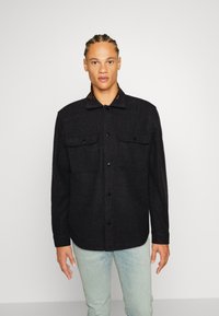 Sort uldskjorte med knapper, der har to forlommer og et mønstret krave, iført med lyseblå jeans. Glat tekstur og afslappet pasform.