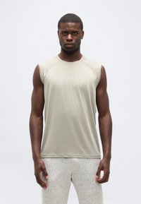 Νεαρός άνδρας με σκουρόχρωμη επιδερμίδα που φοράει αμάνικη μπεζ αθλητική μπλούζα και ανοιχτό γκρι φόρμα, στέκεται μπροστά σε ένα απλό λευκό φόντο.