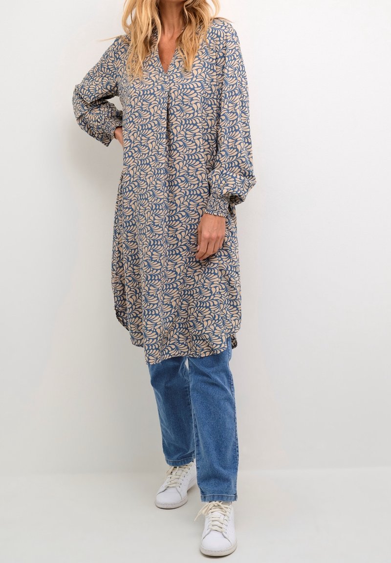 Femme portant une tunique à manches longues, longueur genoux, à motifs, par-dessus un jean bleu et des baskets blanches, debout sur un fond blanc uni.
