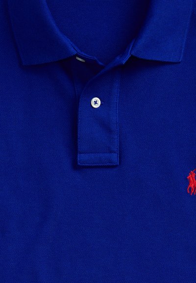 Kék galléros póló, felhajtott gallérral, egy fehér gombbal begombolva, és egy kis piros hímzett logó a jobb oldalon.
