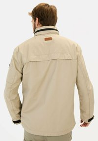 Beżowa kurtka z wysokim kołnierzem i czarnym paskiem. Posiada skórzaną metkę z tyłu oraz regulowane mankiety. Wykonana z lekkiego materiału.