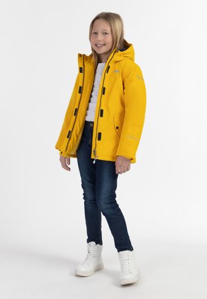 Gul vandtæt jakke med sorte knapper og grå foer, med hætte og lommer, parret med blå jeans og hvide sneakers.