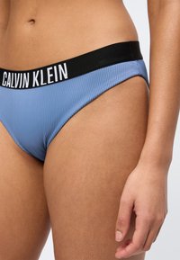 Zbliżenie osoby noszącej prążkowaną niebieską bieliznę Calvin Klein z czarnym paskiem w pasie i białym napisem z logo, z ręką swobodnie opuszczoną wzdłuż ciała.