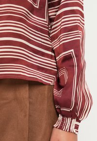Langärmlige Bluse aus burgunderfarbener Seide mit weißen Streifen, ausgestattet mit einem gerafften Bund und einem verkürzten Saum, kombiniert mit braunen Hosen.