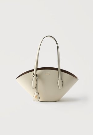 Geantă de mână din piele crem cu formă trapezoidală largă, cu două bretele pentru umăr și etichetă de logo aurie pe un fundal simplu și deschis.