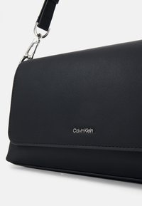 Sac à main en cuir noir avec un fermoir à rabat, présentant un accent en logo argenté et une surface texturée. Comprend une bandoulière amovible.