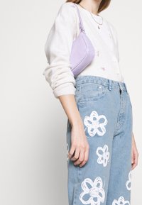 Cardigan chiaro in crema con ricami floreali, abbinato a jeans in denim blu con motivi di fiori bianchi. Tieni in mano una piccola borsa lavanda.