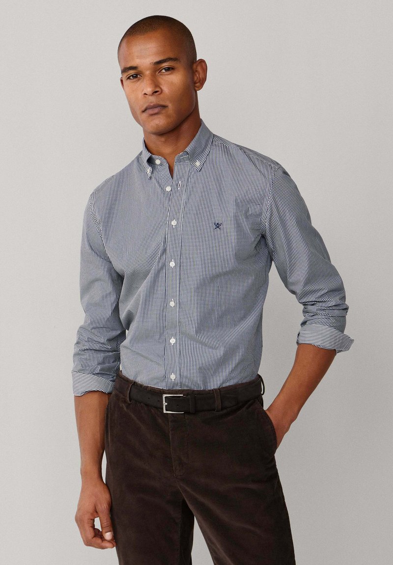 Camisa abotonada con patrón de cuadros en azul marino y blanco, material de algodón, con cuello abotonado y mangas remangadas. Cinturón negro y pantalones marrones.