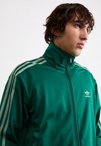 Zelena jakna na zip od glatkog materijala, s tri bijele pruge na svakoj ramenu i izvezenim adidas logotipom na prsima.