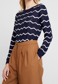 Pull bleu marine avec motif zigzag blanc, poignets côtelés et encolure ronde, associé à un pantalon marron taille haute.