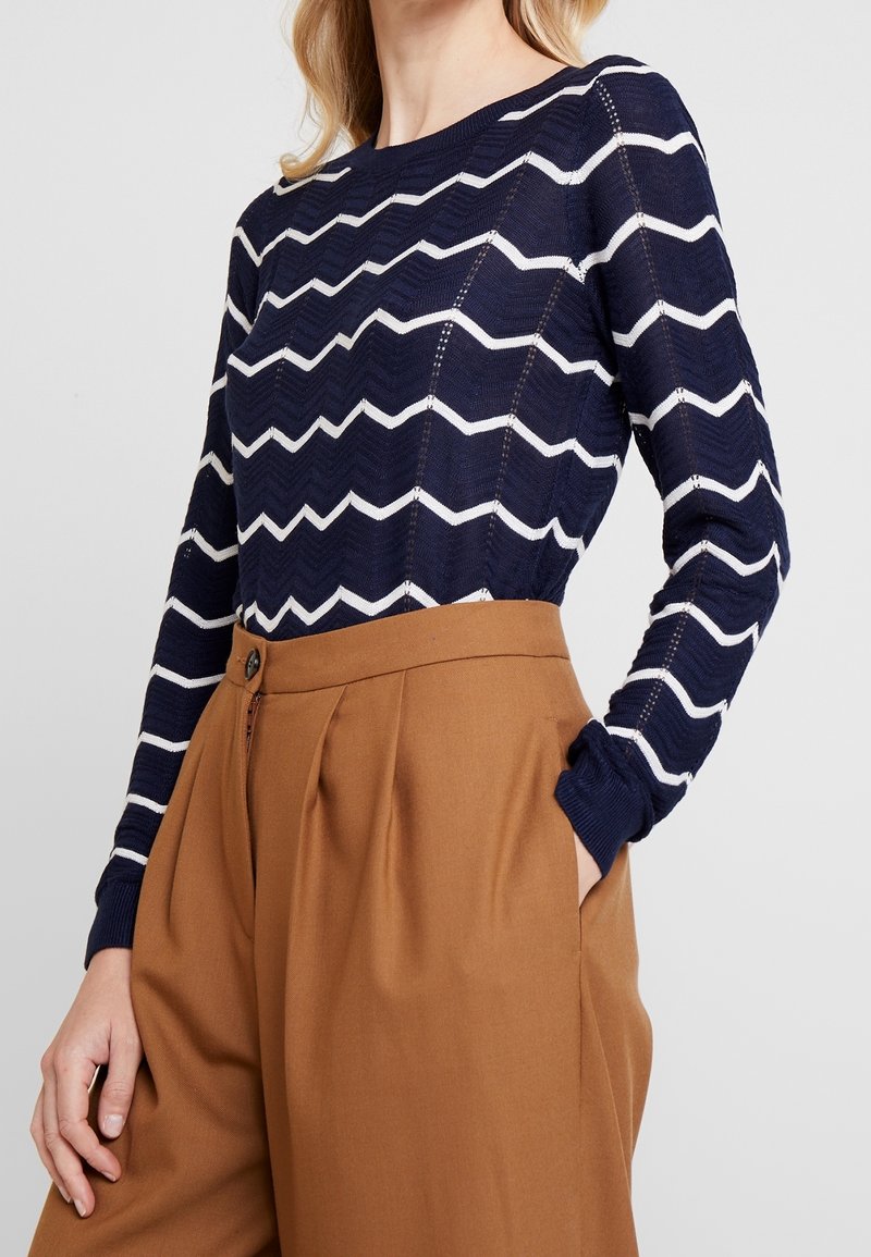 Pull bleu marine avec motif zigzag blanc, poignets côtelés et encolure ronde, associé à un pantalon marron taille haute.