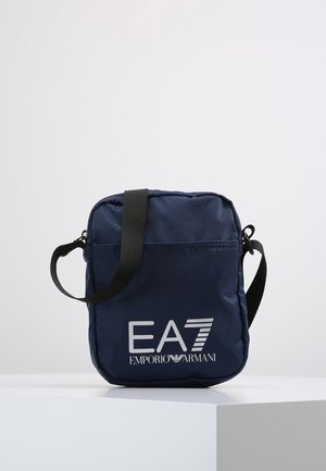 Marineblauwe Emporio Armani crossbody tas met zwarte verstelbare riem en wit EA7-logo aan de voorkant, geplaatst op een wit oppervlak.