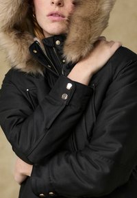 Veste noire avec une capuche doublée de fourrure, comportant des éléments en métal doré et des boutons-pression. Le tissu semble lisse et durable avec une coupe ajustée.